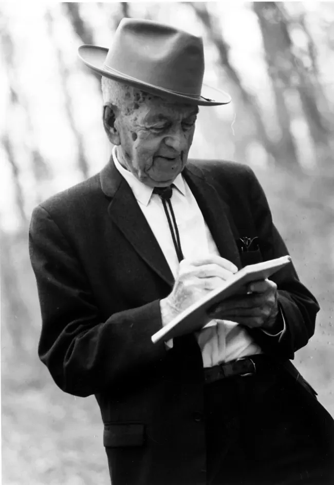 Black-and-white photograph of an elderly Freeman Tilden in a hat and suit, writing in a small notebook outdoors